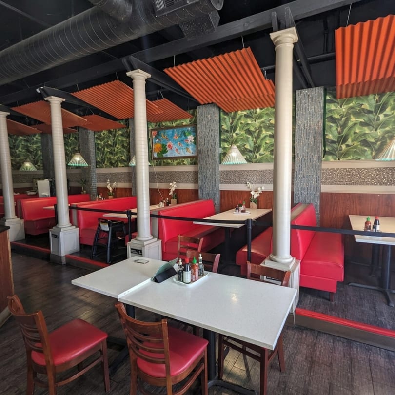 Restaurant interior with red booths, white tables, and tropical leaf wallpaper, under exposed black ductwork.