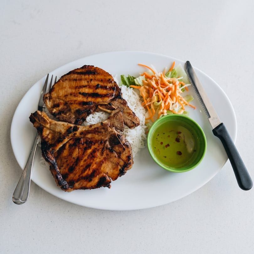 Grilled pork chops with rice, shredded salad, and a green bowl of sauce on a white plate.