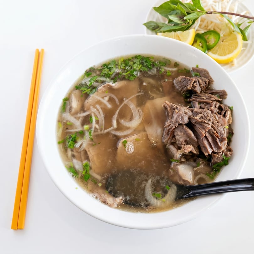 A bowl of pho with beef, noodles, and broth, served with a side of herbs and lime.